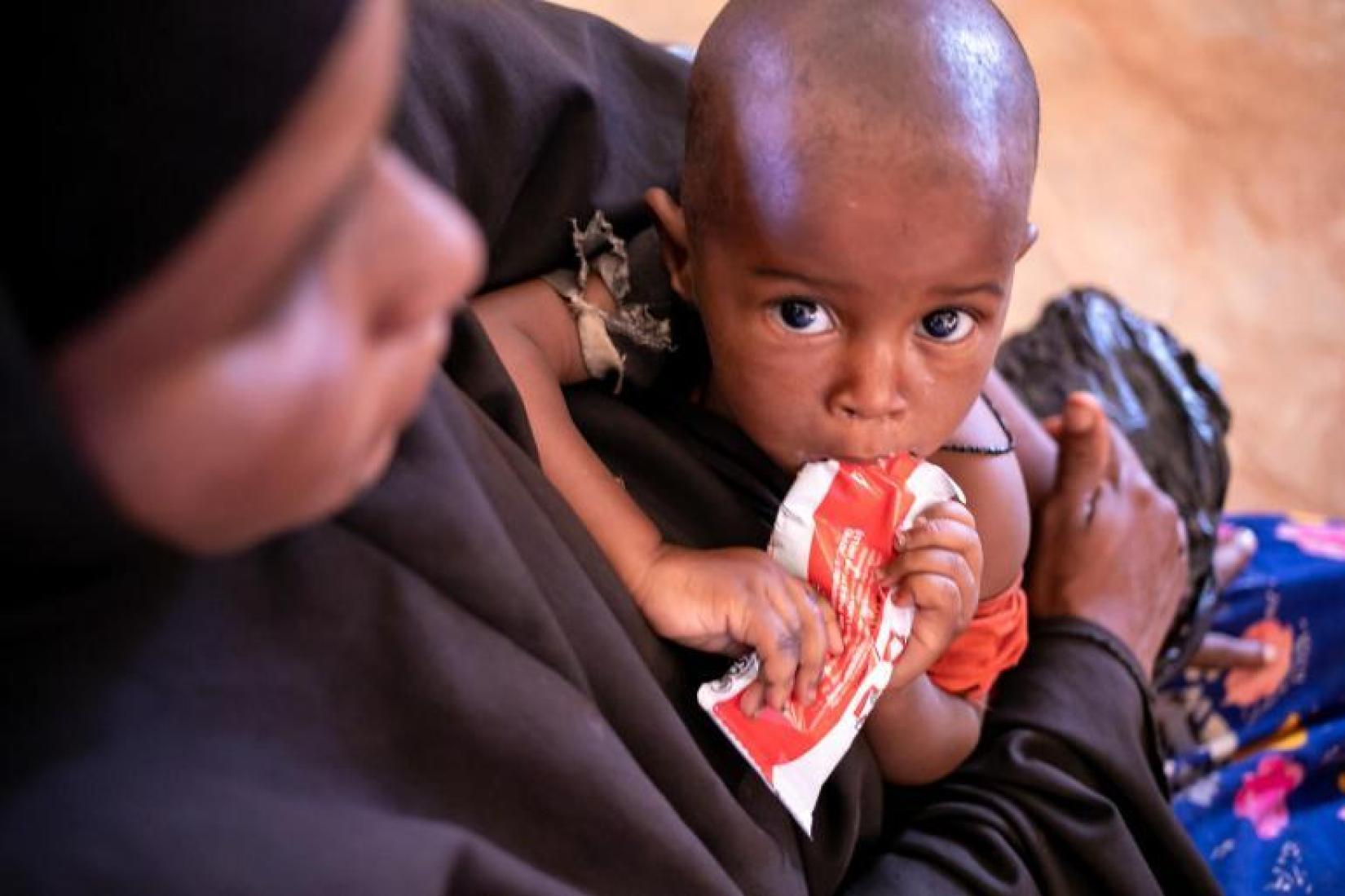 Na Somália, uma criança pequena está no colo de sua mãe enquanto se alimenta com um sachê de alimento terapêutico pronto para uso .