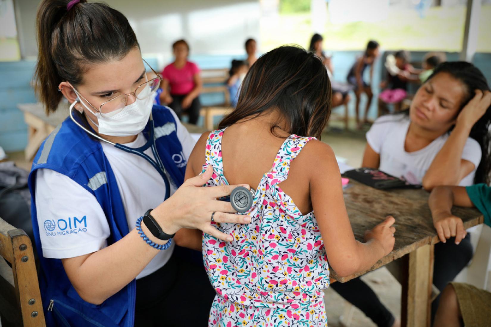 Maria Chan, médica da OIM, auscultando uma menina indígena em Roraima, em fevereiro de 2023. A OIM apoia a assistência médica aos indígenas venezuelanos e às comunidades anfitriãs nas áreas remotas da fronteira entre o Brasil e a Venezuela. 