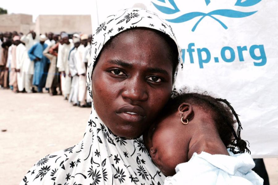 mulher nigeriana segura criança no colo ao lado de fila do programa mundial de alimentos