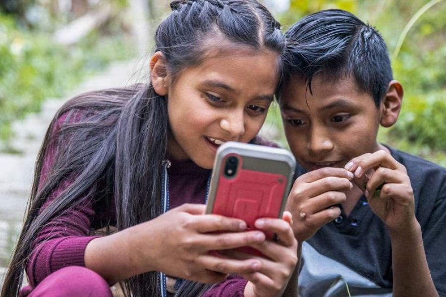Duas crianças brincam ao ar livre enquanto seus pais participam de um workshop sobre segurança online e práticas parentais positivas na Guatemala.